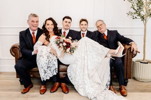 Wedding party posing on leather sofa, groomsmen in navy suits with orange ties, bride holding red bouquet.
