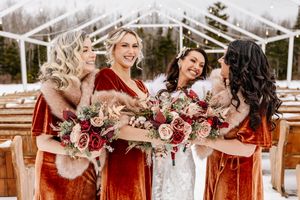 Bridesmaids in rust velvet dresses with fur wraps holding red and pink bouquets at winter outdoor ceremony.