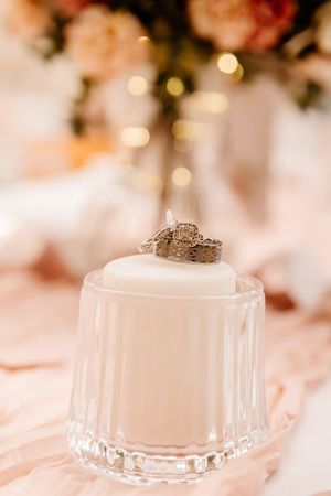 Wedding rings resting on top of a white pillar candle in a crystal glass holder with floral decor.