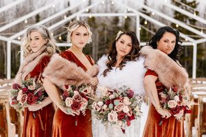 Bride with three bridesmaids in burnt orange velvet dresses and fur shawls posing at snowy winter wedding.