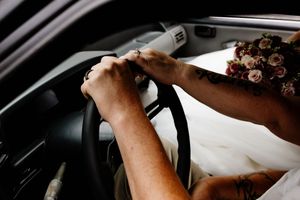Bride and groom holding hands on steering wheel in car, bride wearing white dress with floral bouquet visible.