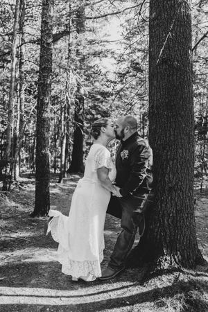 Black and white photo of bride and groom sharing a tender kiss among tall pine trees in a forest.