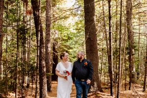Bride and groom walk hand in hand through a sunlit forest during their outdoor wedding ceremony.