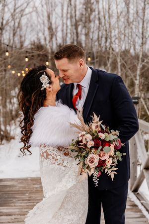 Bride and groom sharing romantic moment on snowy winter bridge, bride in lace dress with fur wrap and bouquet.