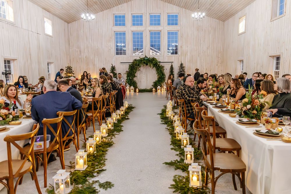 Wedding guests dining at candlelit tables lined with greenery in a bright white barn venue.