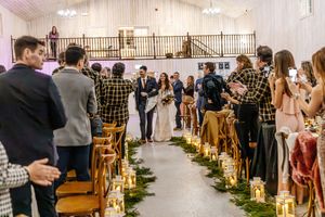 Wedding guests applaud as bride and groom walk down aisle lined with candles and greenery in elegant white barn venue.