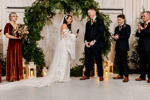Bride in lace gown and groom in navy suit share vows at altar adorned with lush greenery arch and lanterns.