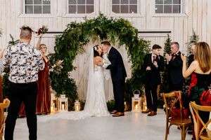Newlyweds share first kiss under stunning circular greenery arch with lanterns in a bright white barn ceremony.