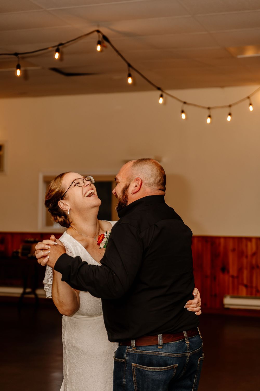 Bride laughs joyfully while sharing a first dance with her groom under warm string lights at the reception.