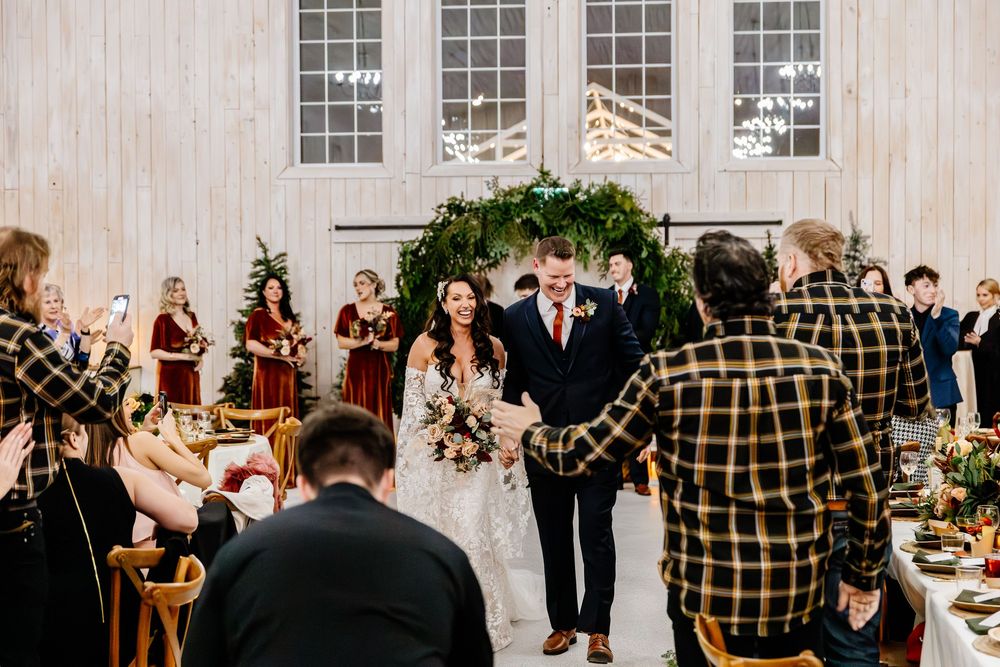 Smiling bride and groom walk back down aisle after wedding ceremony in white barn with greenery arch backdrop.