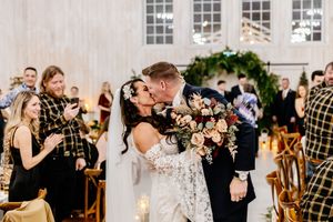 Bride and groom share romantic kiss in aisle surrounded by guests celebrating with lush floral bouquet in hand.
