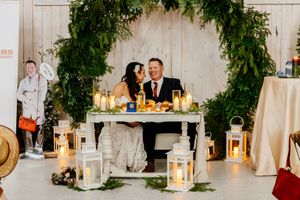 Newlyweds sit together at sweetheart table surrounded by white lanterns and lush greenery arch at reception.
