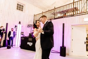 Bride and groom share first dance in elegant white venue with purple uplighting and DJ booth in background.