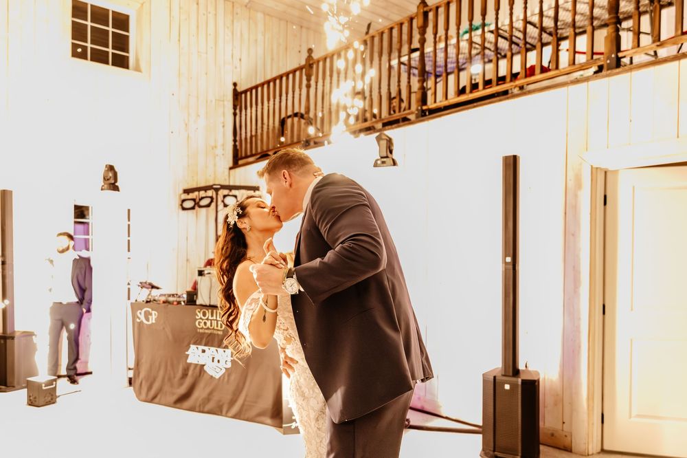 Bride and groom share romantic first dance kiss in warmly lit barn reception venue with wooden staircase.