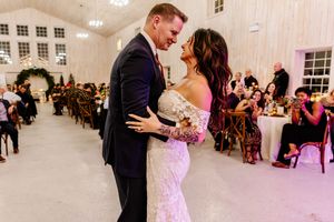 Bride in lace gown and groom in navy suit share intimate first dance in white barn reception hall.