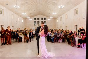 Bride and groom share first dance in elegant white barn wedding venue with chandeliers and seated guests.