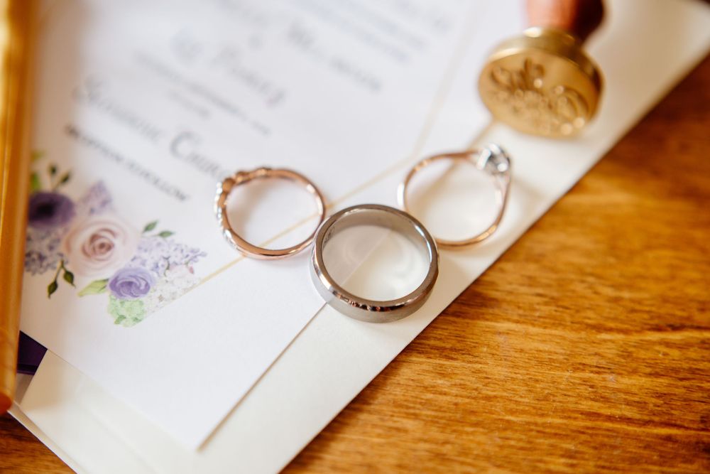 Three wedding rings, including diamond engagement ring, displayed on floral wedding invitation with wax seal.