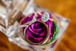 Diamond oval engagement ring with twisted band resting on gold wedding band beside purple rose decoration.