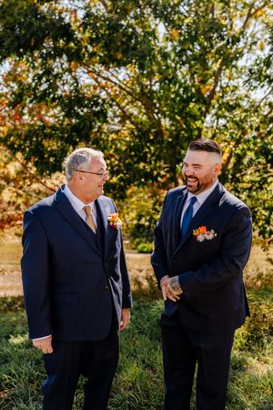 Two men in navy suits with autumn boutonnieres laugh together outdoors at a fall wedding.