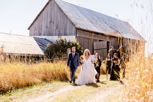 Bride and groom walk with wedding party past a rustic wooden barn through golden autumn fields.