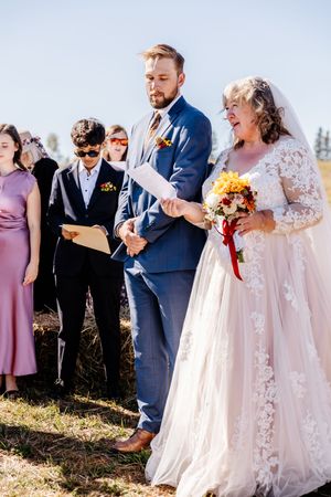 Mature bride in lace gown holding bouquet stands with groom reading vows during outdoor fall wedding.