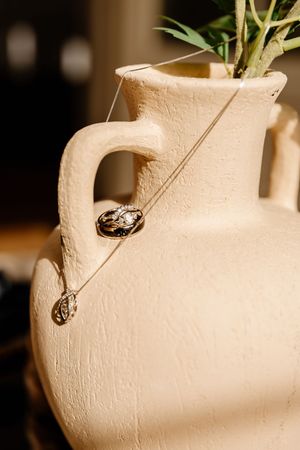 Delicate gold necklace with pendant draped over rustic cream ceramic vase with greenery.
