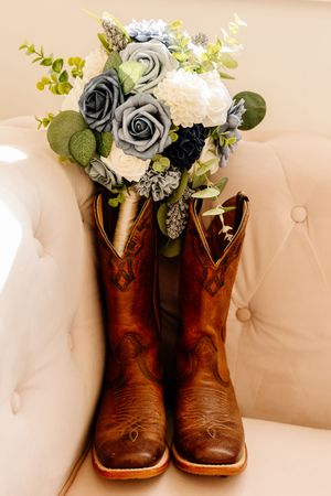 Brown leather cowboy boots holding blue and white rose bridal bouquet on cream tufted chair.