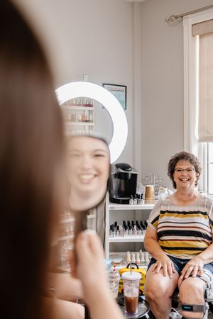 Bride smiles at her reflection while an older woman watches during wedding makeup preparations.