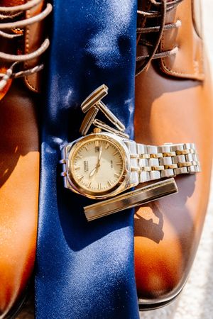 Grooms accessories including brown leather shoes, blue tie, gold watch, and cufflinks arranged for wedding day.