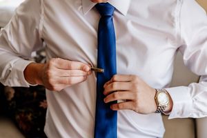 Groom fastens a tie bar on his blue tie while wearing a white shirt and gold watch.