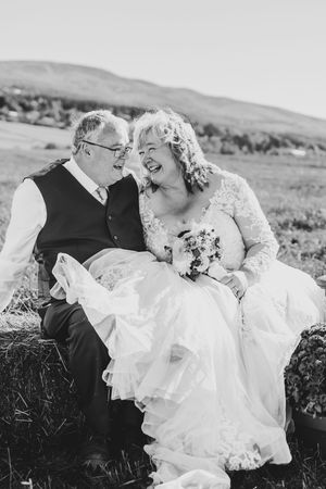 Black and white photo of mature bride and groom laughing together while seated on hay bales in field.