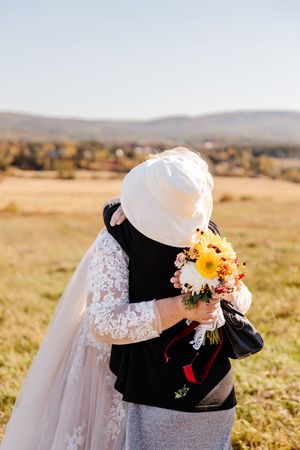 Bride in lace wedding gown embracing elderly guest while holding colorful bouquet in sunny field.