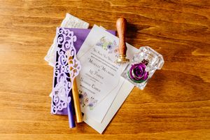 Elegant purple wedding stationery with wax seal, ink bottle, and floral details on wooden surface.