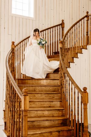 Bride in white gown holding bouquet descending elegant curved wooden staircase in rustic venue.