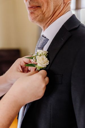 Hands pinning a white rose boutonniere onto a mans dark suit jacket before wedding.