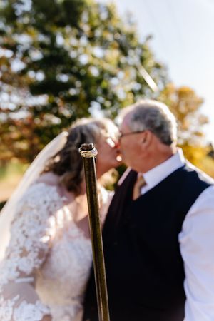 Wedding rings displayed on a pole in foreground as mature couple shares a kiss outdoors in soft focus.