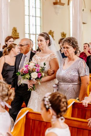 Bride walks down church aisle with parents, holding colorful bouquet of pink and white flowers.
