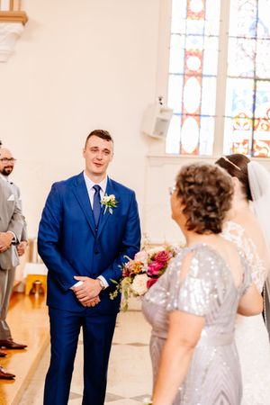 Groom in blue suit watches bride walk down the aisle in a bright church.