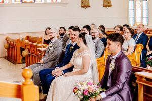 Bride and groom sit together in church pew, smiling during wedding ceremony with guests.