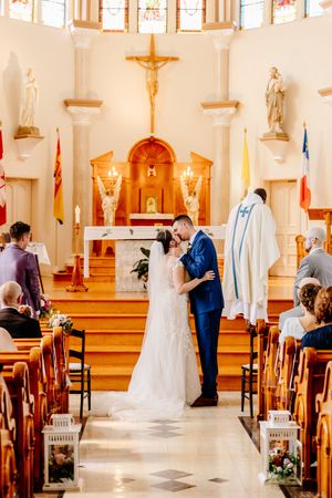 Bride and groom share first kiss at Catholic church altar during wedding ceremony with priest standing behind them.