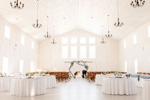 Elegant white wedding reception hall with chandelier lighting, floral arch altar, round tables, and wooden chairs in a barn venue.