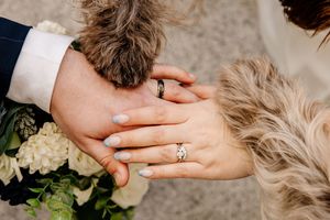 Couple's hands with wedding rings resting on a fluffy dog beside a white floral bouquet.