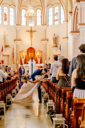 Groom dips bride for romantic kiss in church aisle as wedding guests celebrate around them.
