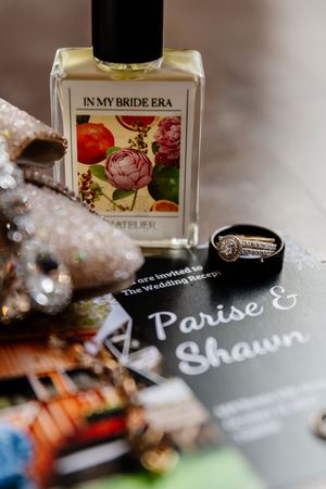 Wedding flat lay featuring In My Bride Era perfume, diamond ring, and Parise and Shawn invitation.