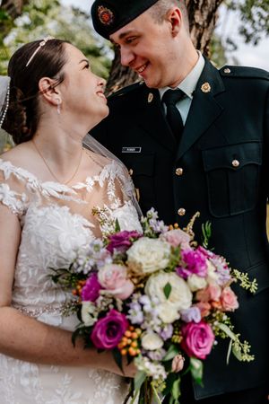 Military groom in Canadian Army dress uniform smiles at bride holding colorful pink and white wedding bouquet.