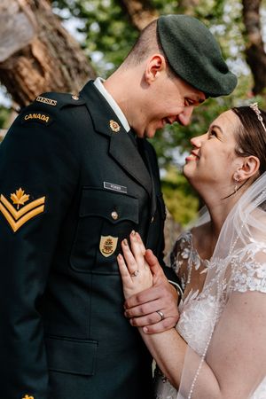 Canadian soldier groom in uniform and bride in lace gown share a tender moment under a tree.
