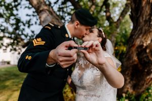 Bride and groom kiss while displaying wedding rings in a small decorative box outdoors.
