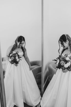 Black and white photo of a bride in a white gown holding a bouquet, reflected in a mirror.