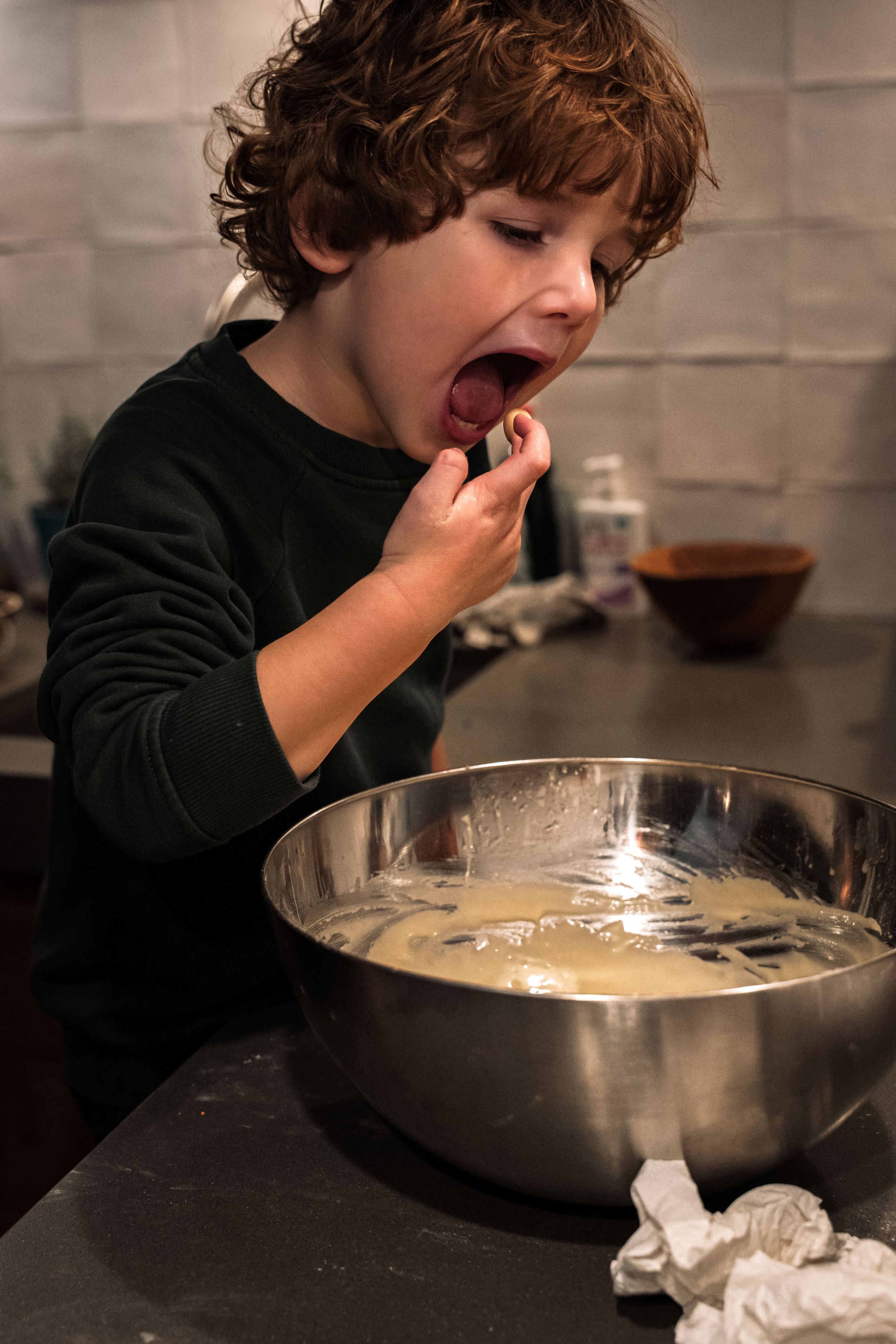 Child in dark sweater tasting ingredients while baking at kitchen counter.