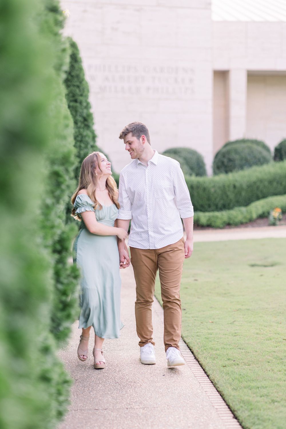 A couple in light summer attire walking together on a paved pathway beside a manicured lawn and hedges.